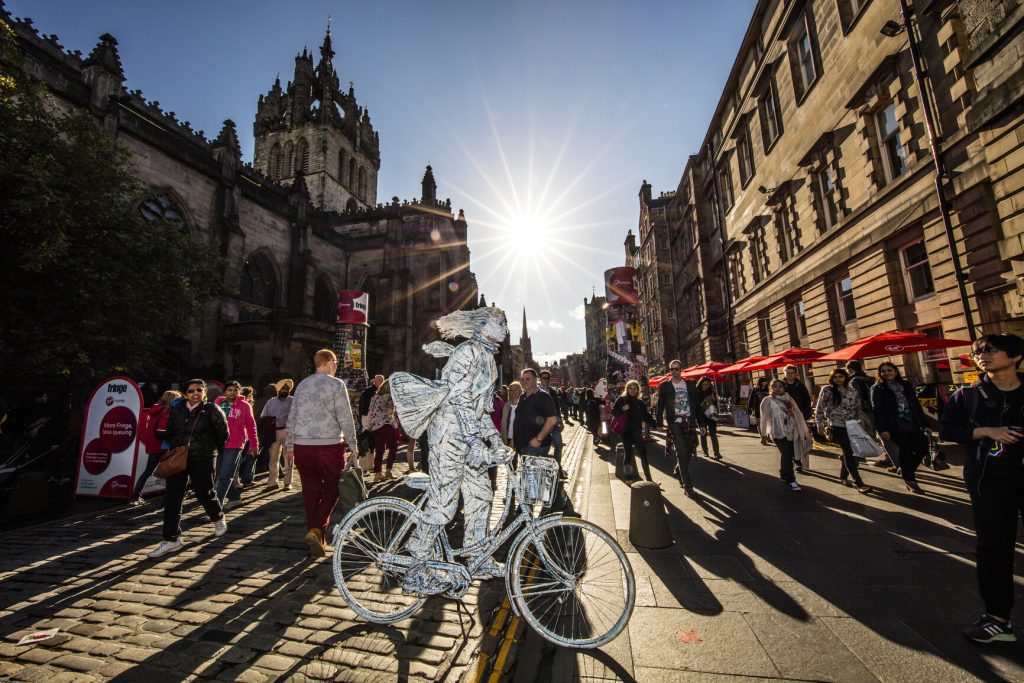 The Edinburgh Festival and Edinburgh fringe festival. A street performer drawing a crowd in a summer performance. A living statue, man in silver on a bicycle in costume. Lothian, Scotland